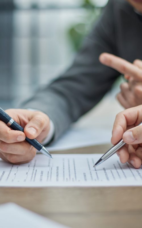 Two professionals review documents at a wooden desk. Hands gesture, pens poised, indicating focused discussion. Natural light filters through a window in the background