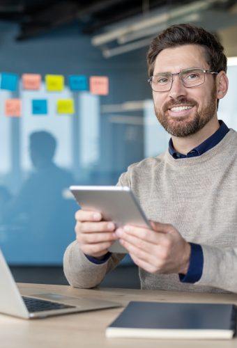Smiling businessman using a digital tablet at his desk in a modern corporate office, representing contemporary work practices, technology integration, and professional success