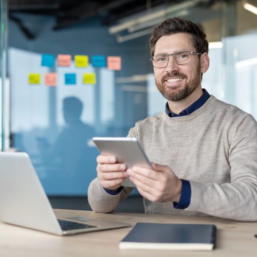 Smiling businessman using a digital tablet at his desk in a modern corporate office, representing contemporary work practices, technology integration, and professional success