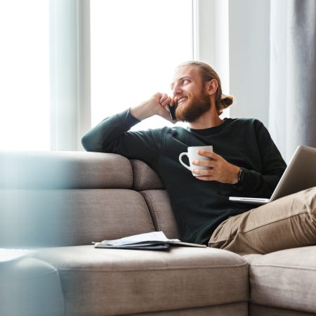 Image of a handsome young bearded man sitting in home using laptop computer talking by phone.