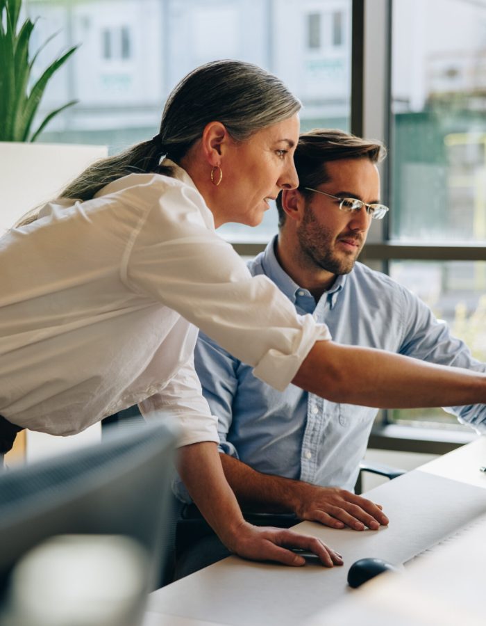 Mature woman working with young man sitting at desk and showing something on computer screen in office. Female executive pointing at desktop monitor and talking with male colleague in office.