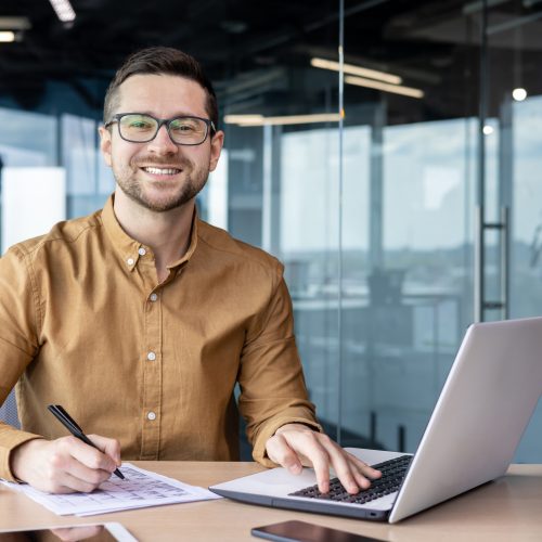 Portrait of young businessman in shirt, man smiling and looking at camera at workplace inside office, accountant with calculator behind paper work signing contracts and financial reports.