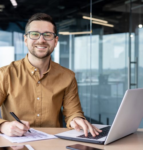 Portrait of young businessman in shirt, man smiling and looking at camera at workplace inside office, accountant with calculator behind paper work signing contracts and financial reports.